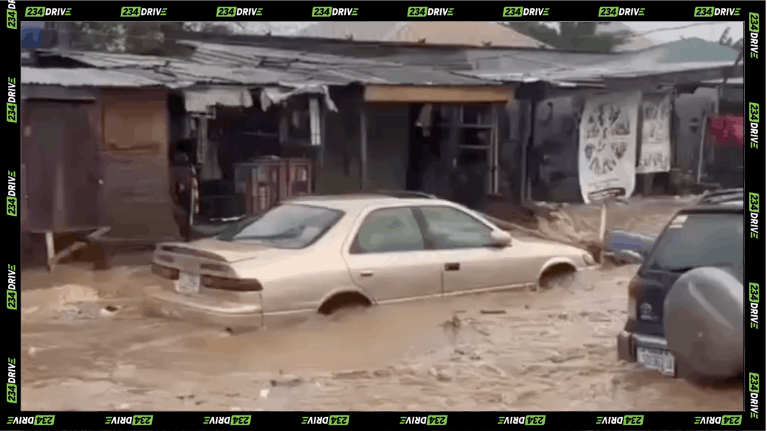A beige sedan sits partly submerged in muddy floodwater outside shops in Shogunle and Mafoluku, Oshodi, after heavy rainfall hit Lagos.