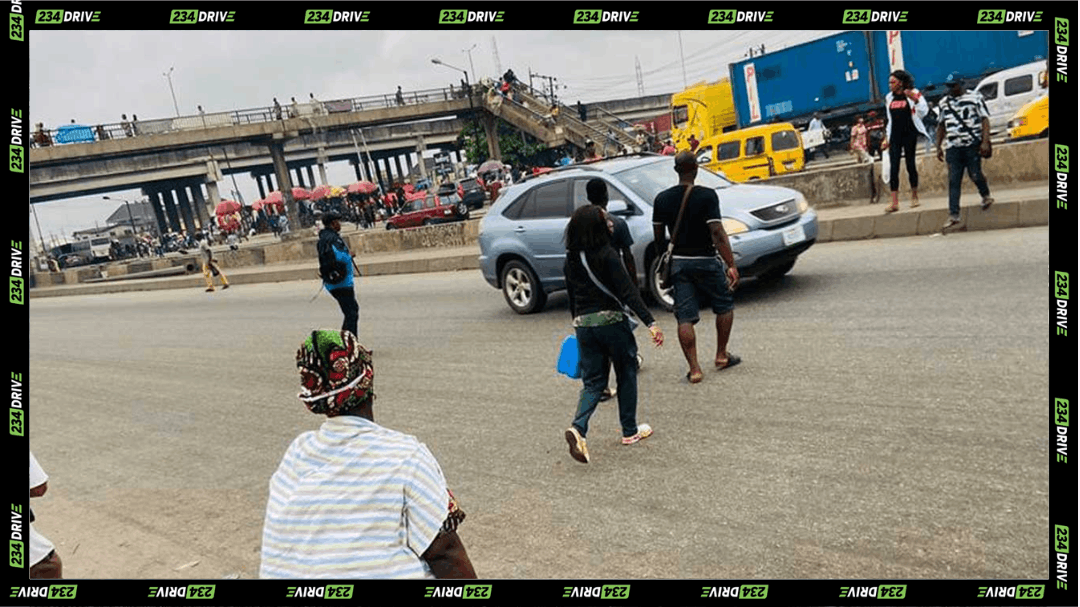 Pedestrians walk across a Lagos expressway as vehicles pass and a pedestrian bridge stands nearby.