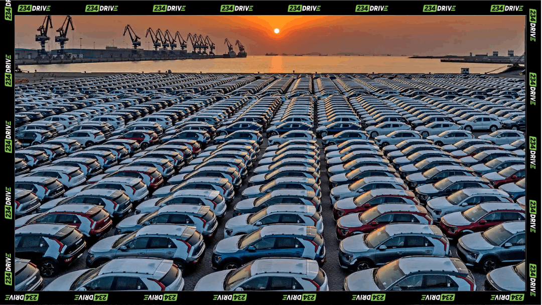 Rows of imported cars parked at a large port terminal, with cranes and a sunset in the background.