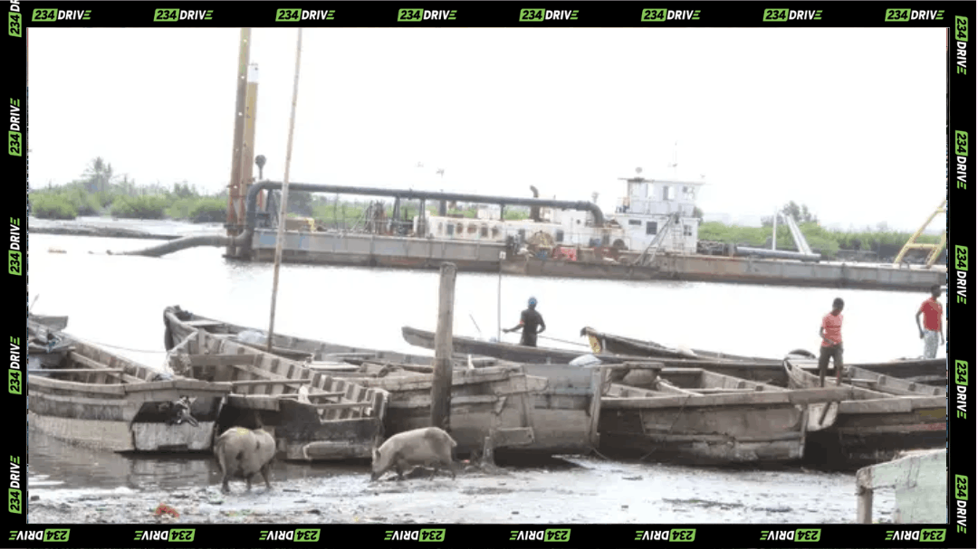 A dredging vessel works on a Lagos lagoon near wooden boats, with workers standing along the muddy shoreline in view.