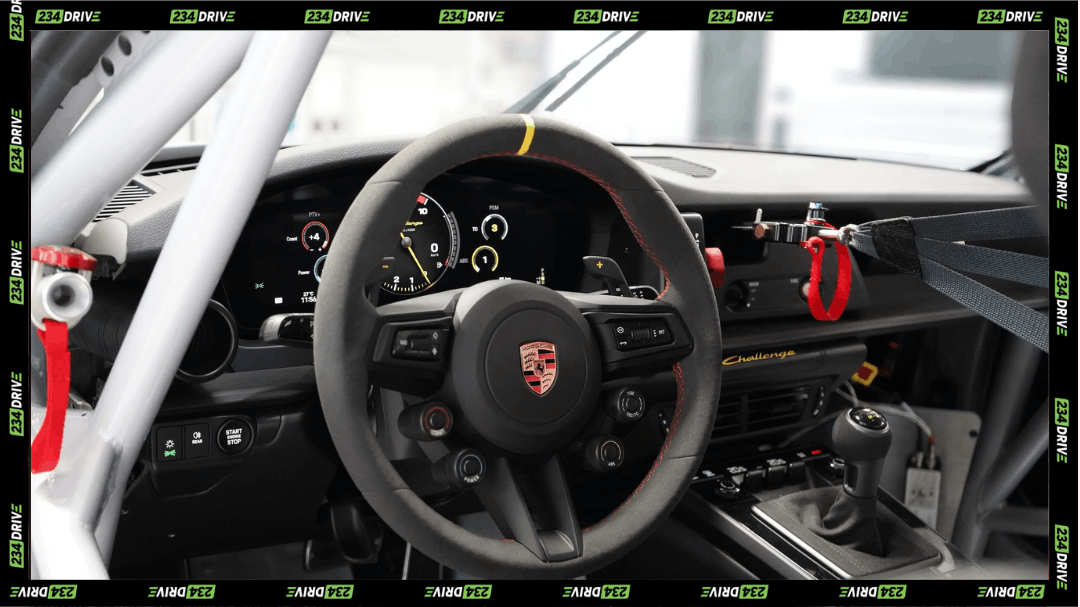 The Porsche 911 Challenge cockpit centres its racing steering wheel, digital display, roll cage, dashboard controls and safety harness for track competition.