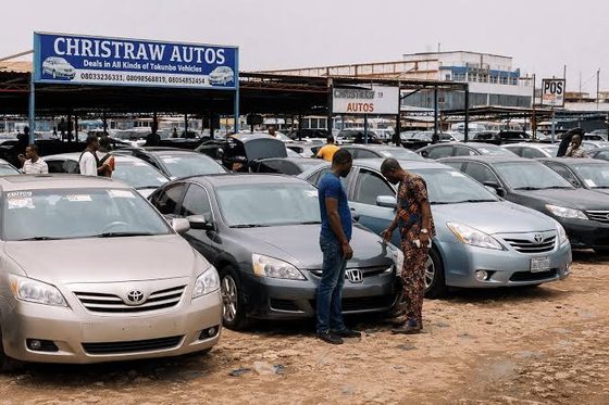 An outdoor car dealership lot in Nigeria filled with rows of imported sedan vehicles under a branded sign for Christraw Autos, where prospective buyers inspect a grey car.