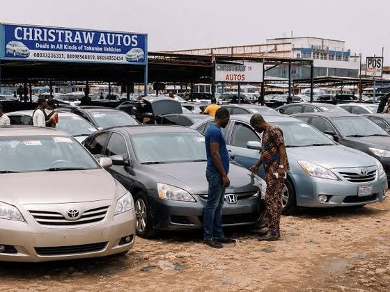 An outdoor car dealership lot in Nigeria filled with rows of imported sedan vehicles under a branded sign for Christraw Autos, where prospective buyers inspect a grey car.