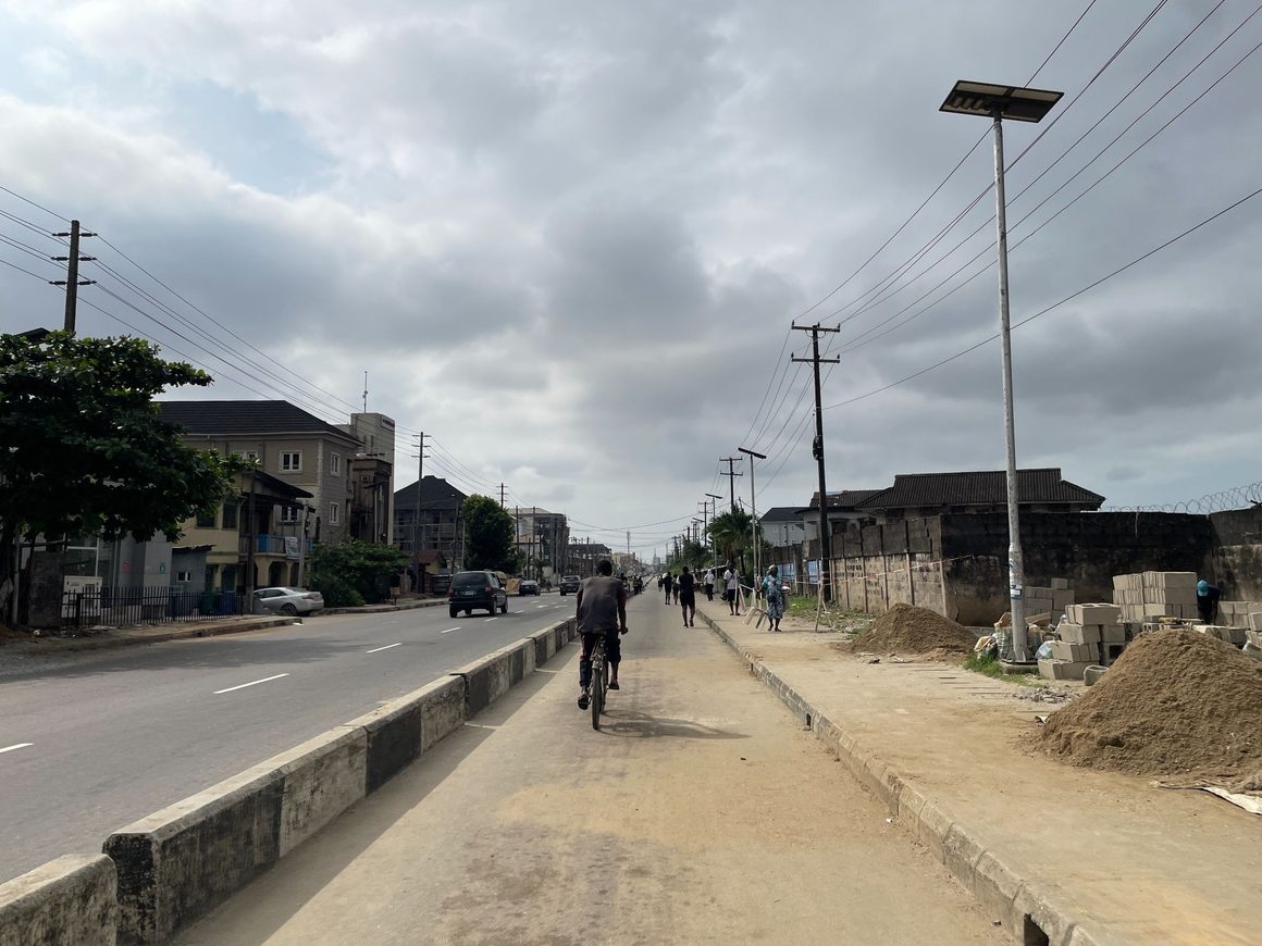 A cyclist and pedestrians travelling along a concrete walkway and road margin next to active vehicular traffic in Lagos.