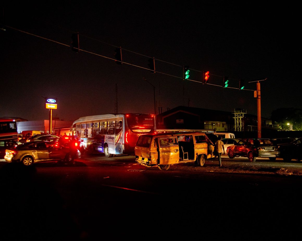 Yellow Danfo bus and vehicles stopped at a dark Lagos intersection beneath overhead traffic lights at night