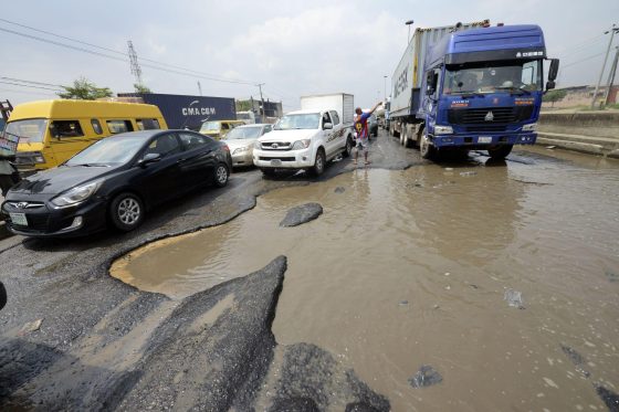 A car drives through standing water and a damaged road in Nigeria, illustrating the challenges motorists face on poorly maintained roads.