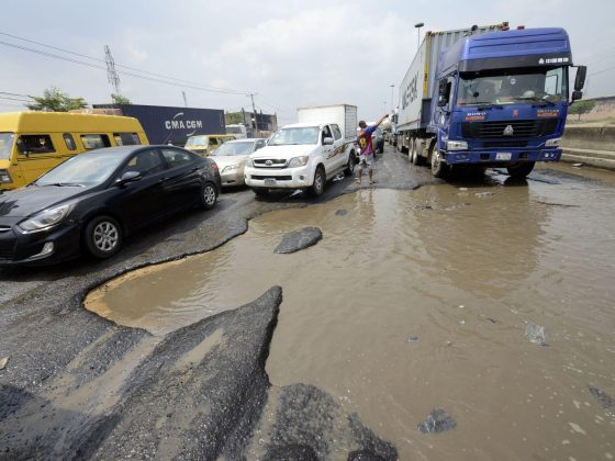 A car drives through standing water and a damaged road in Nigeria, illustrating the challenges motorists face on poorly maintained roads.