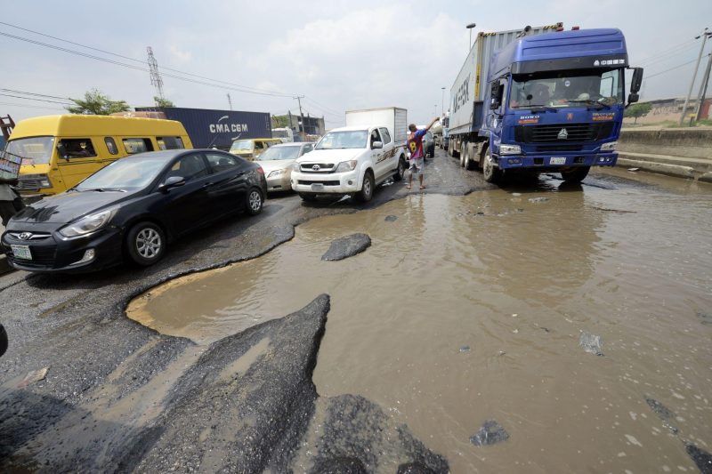 A car drives through standing water and a damaged road in Nigeria, illustrating the challenges motorists face on poorly maintained roads.