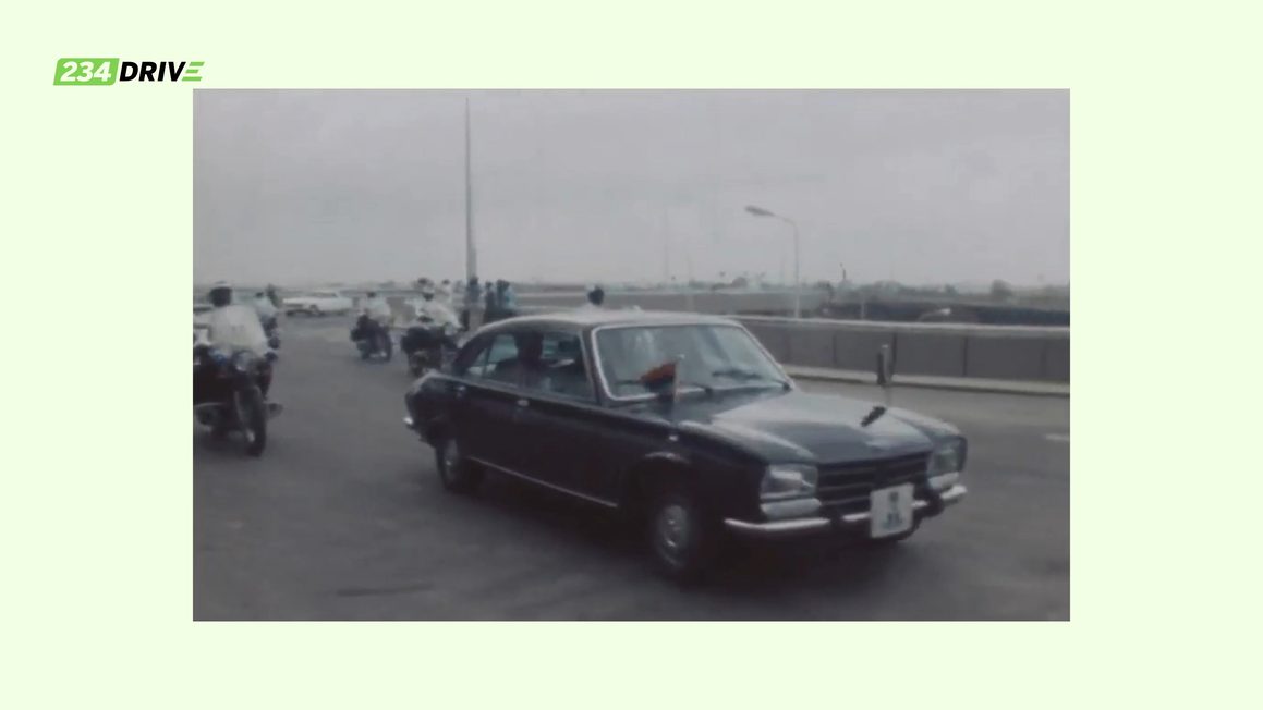 Archive footage of a dark saloon with a flag on the bonnet, escorted by motorcycle outriders